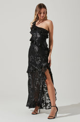 A woman is wearing a black, one-shoulder dress with ruffles and floral lace patterns. The dress features a sheer, flowy lower half with a high slit revealing her leg. She has on black high-heeled sandals with ankle straps.